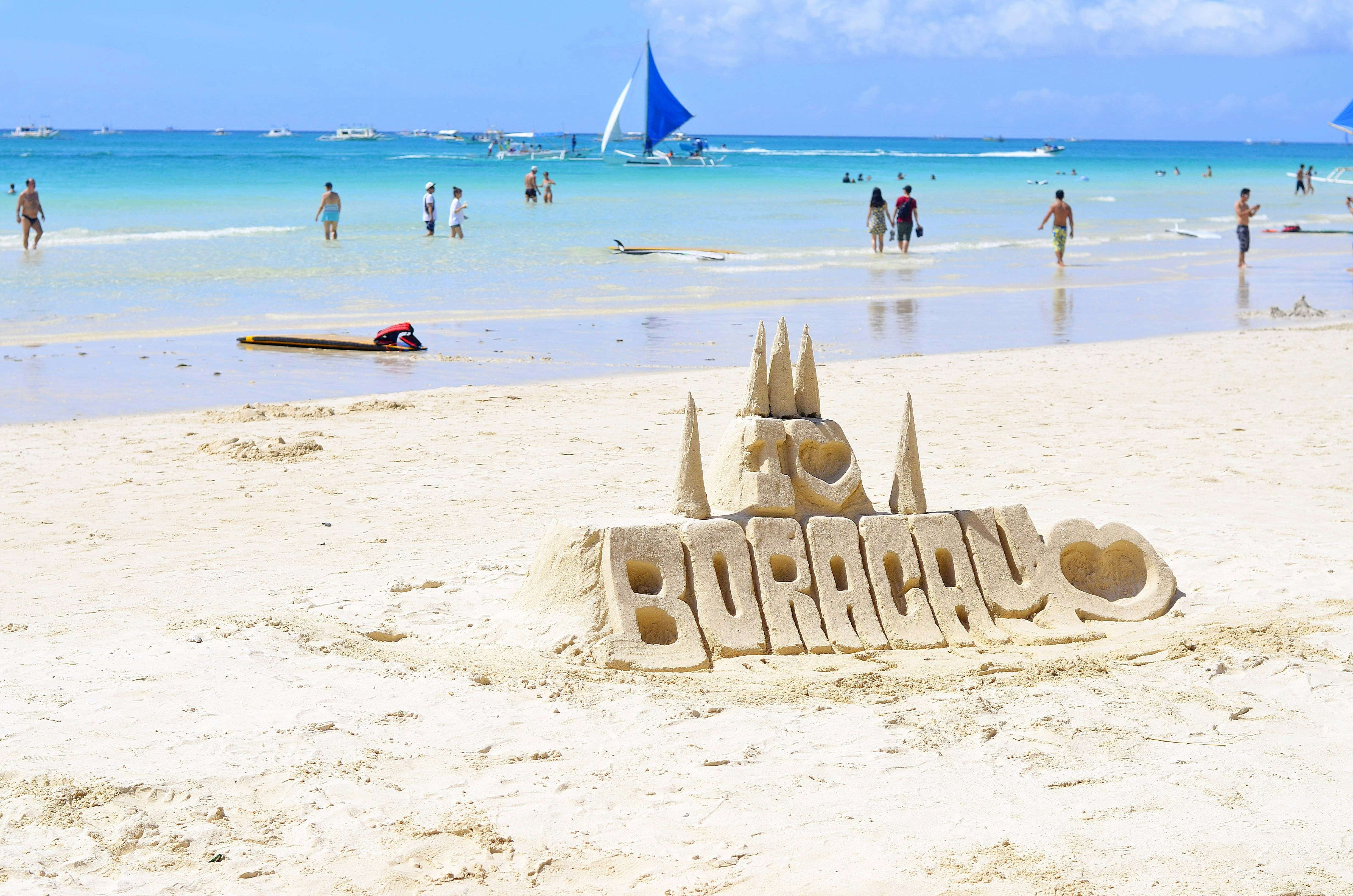 A large sand sculpture on a bright white-sand beach spelling out 'I [heart] BORACAY', topped with small castle spires. In the background, people are wading and swimming in clear turquoise water under a sunny blue sky, with a distinctive blue sailboat sailing in the distance.