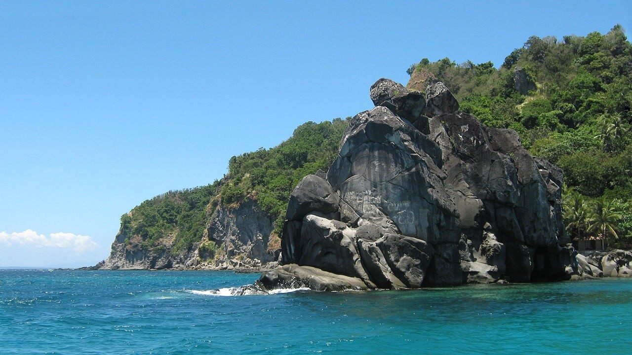 Rocky islet rising from clear turquoise water off the coast of Negros Oriental Philippines with lush green cliffs behind