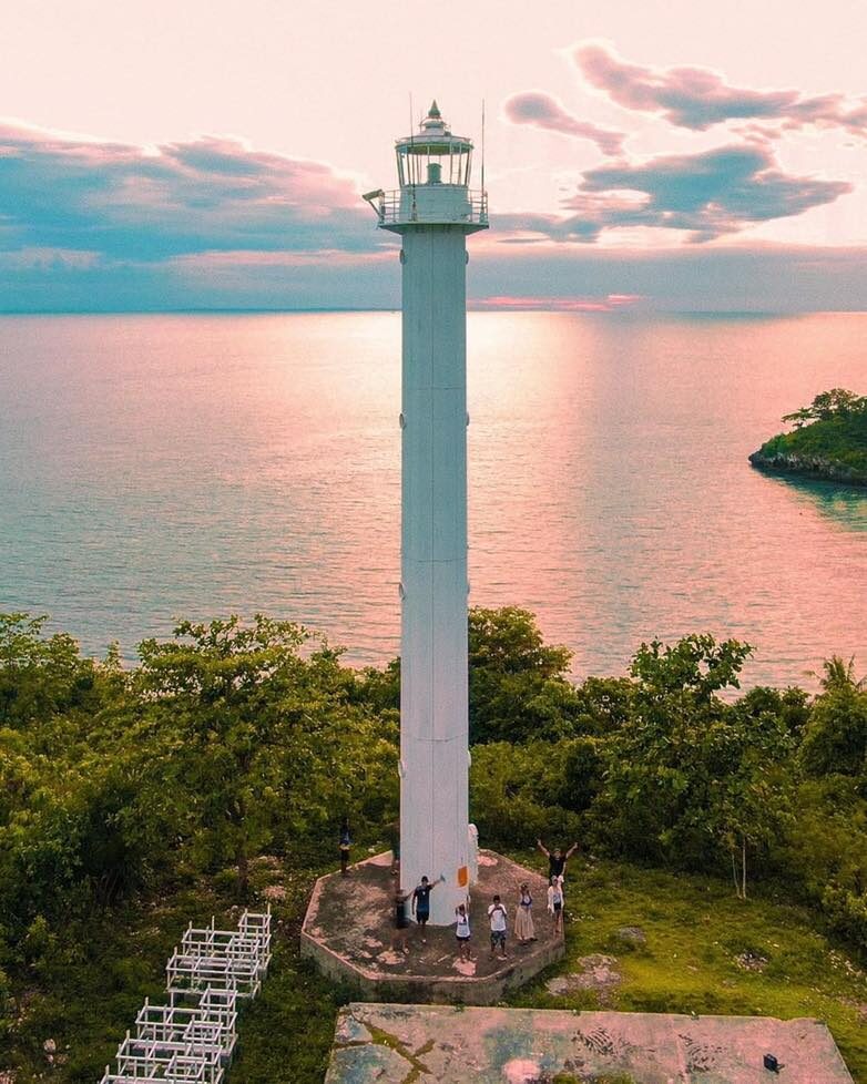 Malapascua Lighthouse tall white tower at sunset with pink-purple sky reflected in the calm sea and visitors at the base