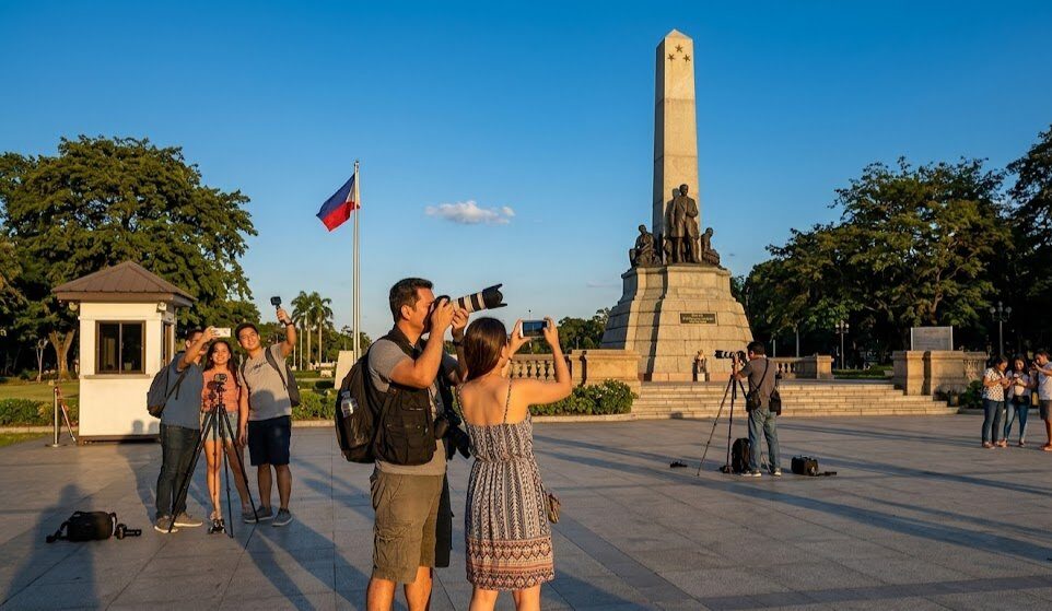 Photographers at Rizal Park Manila Luneta with the Rizal Monument obelisk and Philippine flag at sunset
